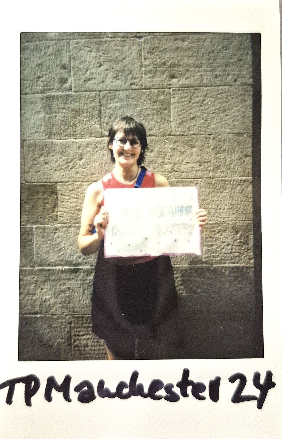 A polaroid of a smiling person holding a colorful sign stands against a stone wall, with text below reading "TP Manchester 24"