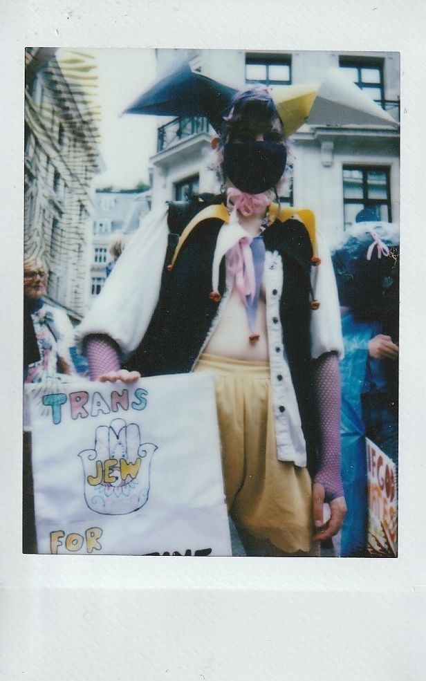 A person in a colorful outfit holds a sign reading "Trans Jew for Palestine" at a public event.