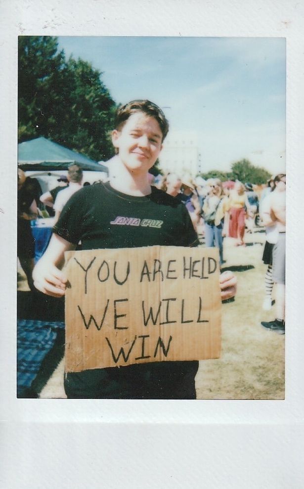 A person is holding a sign reading "YOU ARE HELD WE WILL WIN".