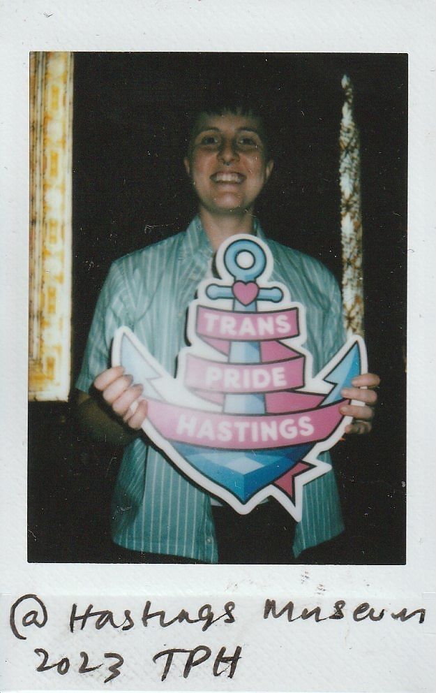 A person smiles and holds a colorful sign reading “Trans Pride Hastings” at Hastings Museum.