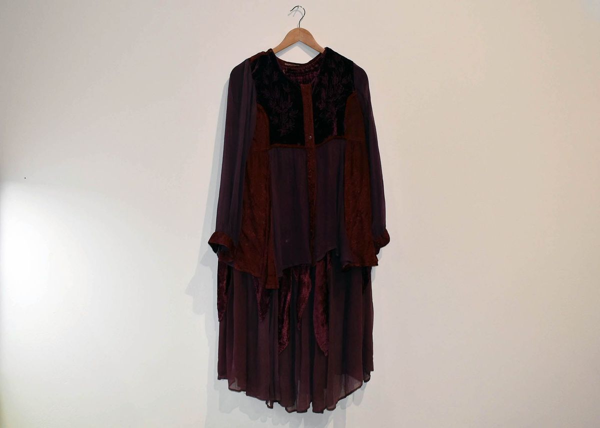 A long-sleeved, dark purple dress hangs on a wooden hanger against a plain, white wall.
