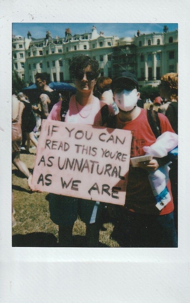 Two individuals at a public event display a sign stating, "If you can read this, you're as unnatural as we are".
