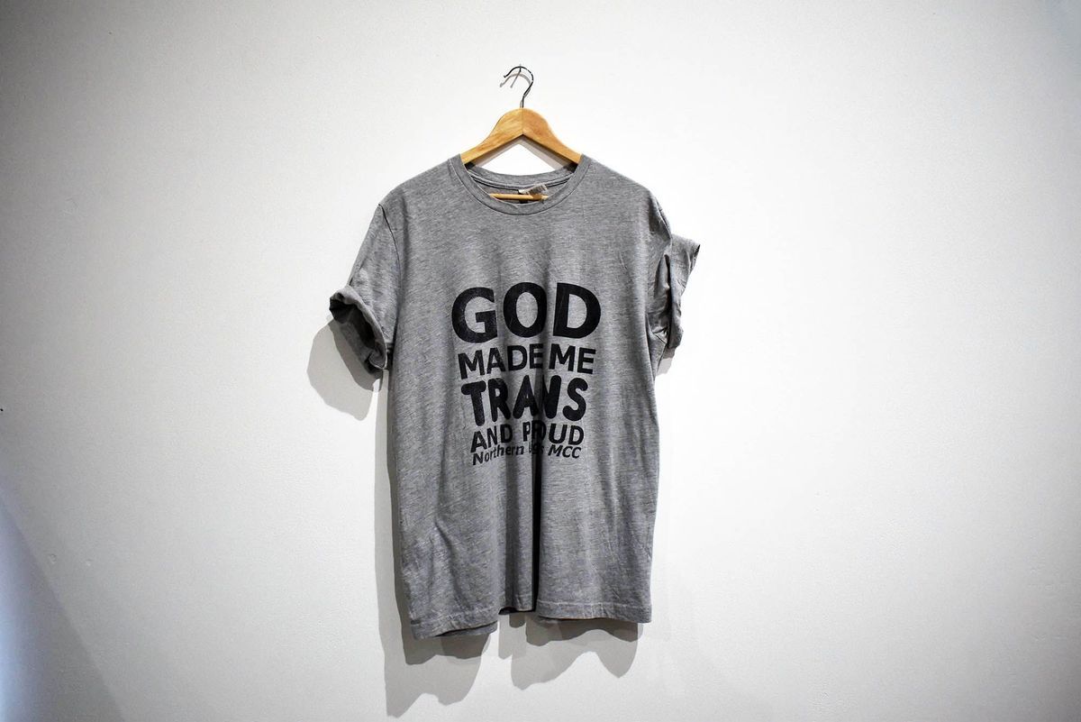 A grey t-shirt on a wooden hanger with text "GOD MADE ME TRANS AND PROUD Northern Lights MCC"