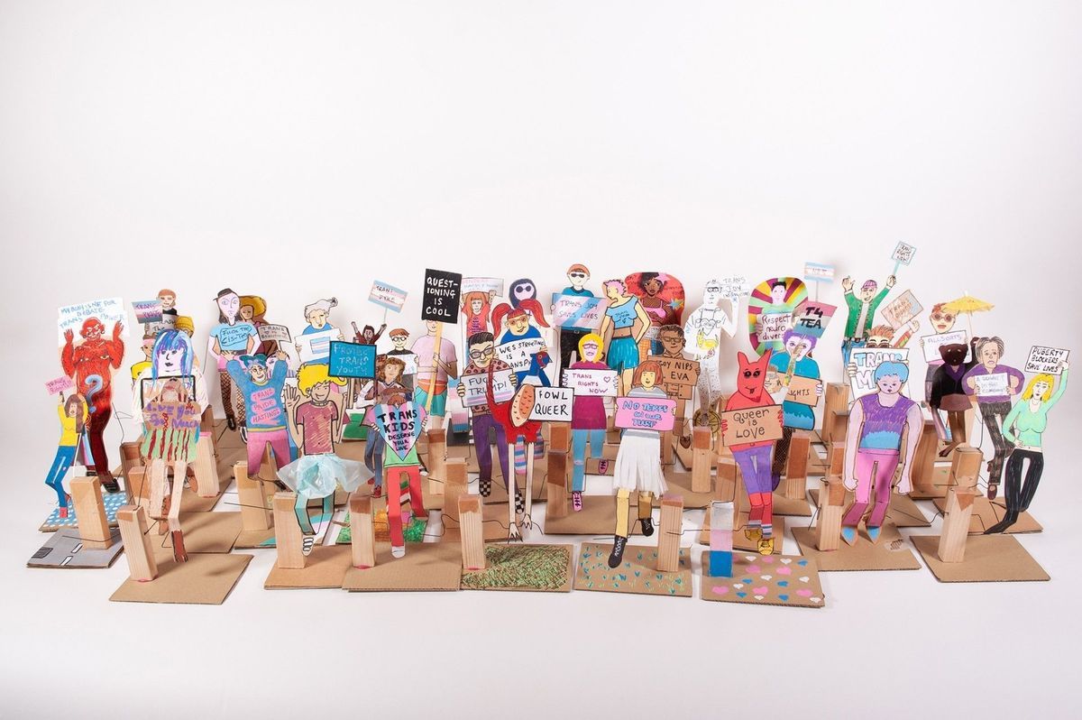A photograph which is on a white background and has a large group of 'paper people' which are made from cardboard. The people are colourful, wearing different outfits and an array of expression. All of them are holding placards which say different phrases such as 'Trans Kids Deserve Your Love' and 'Queer Love'.