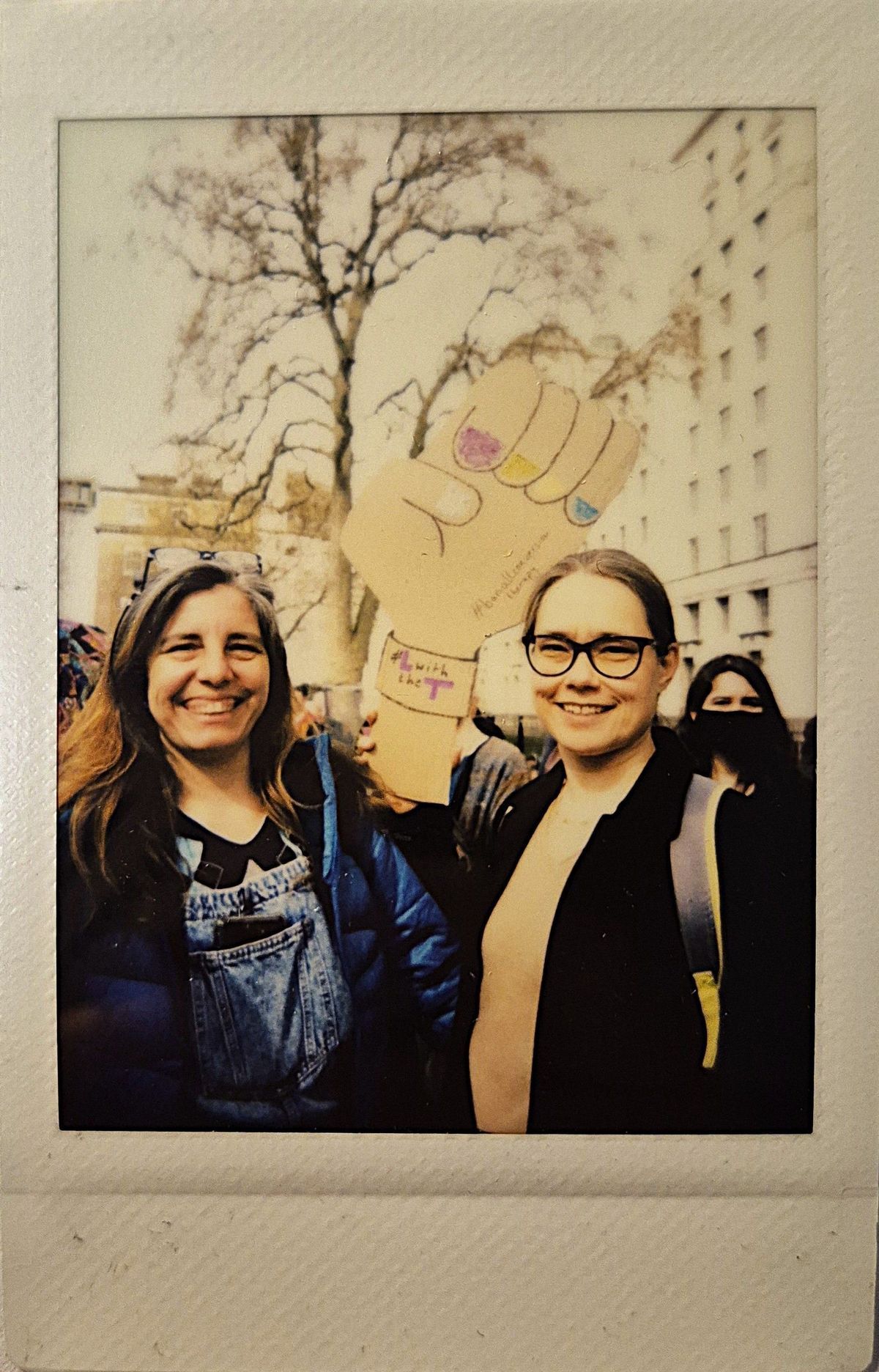 Two smiling individuals hold a paper fist sign with colorful nails in a park during a public event.