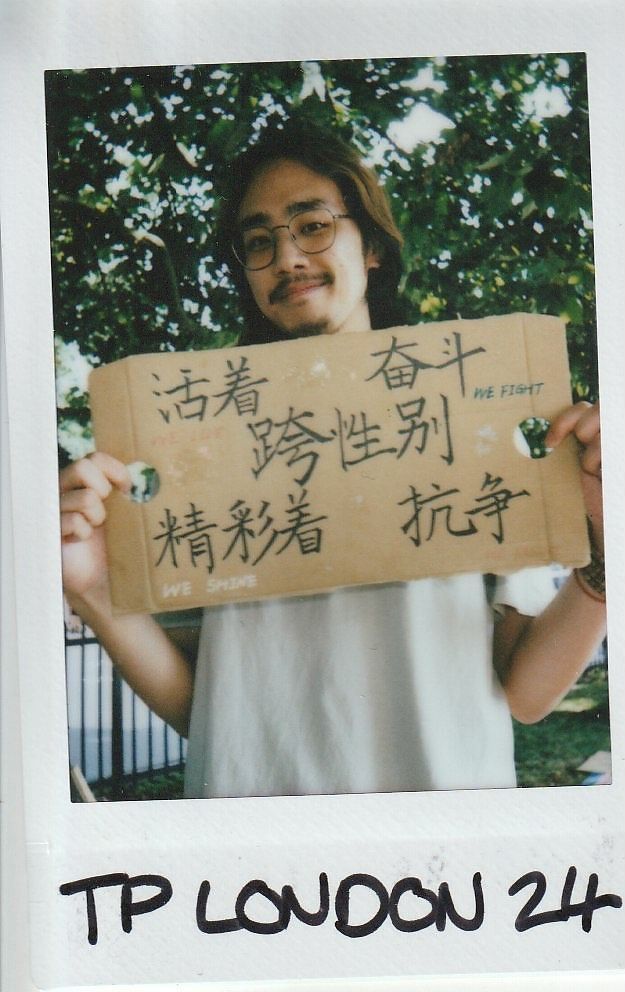 A person holding a cardboard sign with handwritten text, standing outdoors near green leaves.