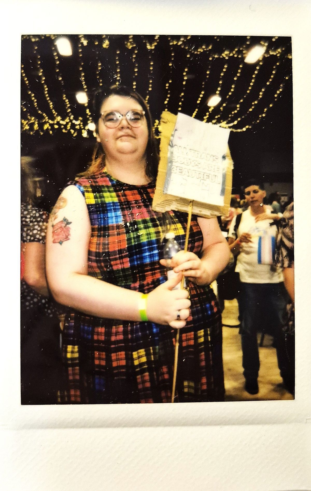A person in a colorful plaid dress holds a sign in a festive, string-lit venue.