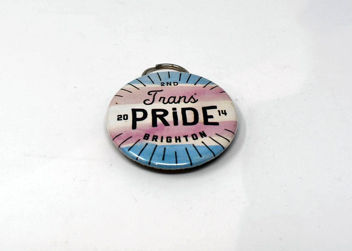 Bottle opener, metal, made to look like a badge reading "2nd Trans Pride Brighton 2014". Blue, white, pink with attached ring on reverse.