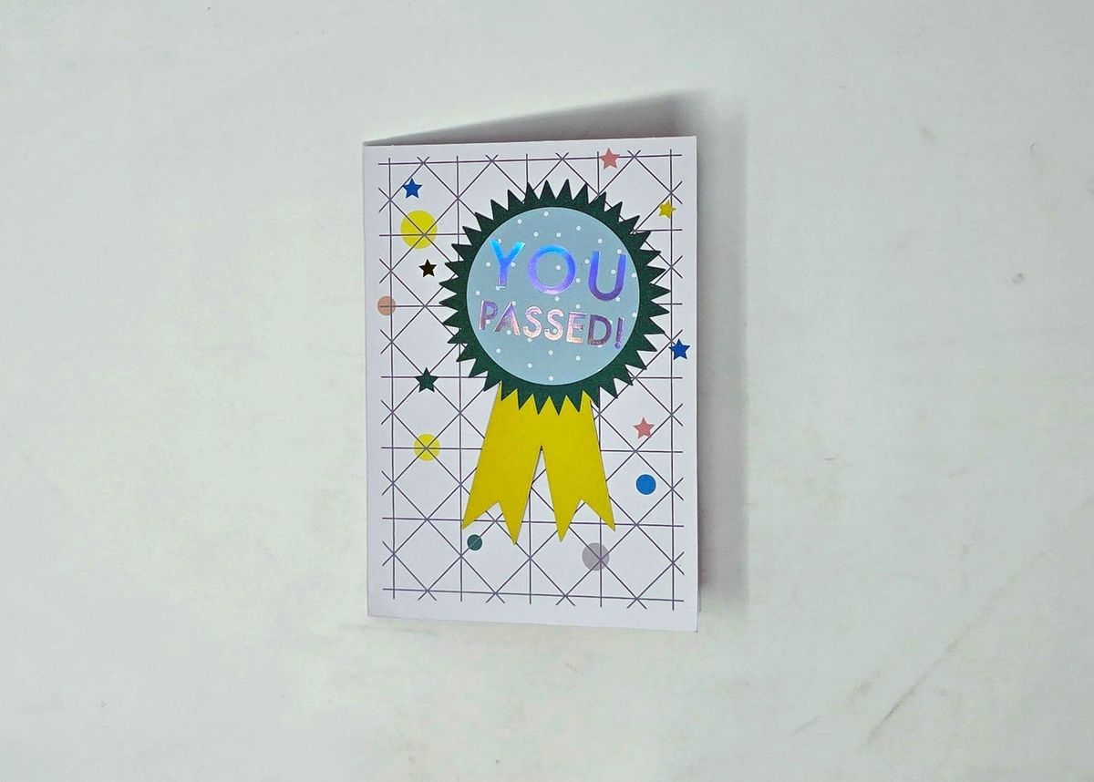 The greetings card that says "YOU PASSED!" inside a blue and green rosette with a yellow ribbon on a background of geometric lines and coloured stars and circles.