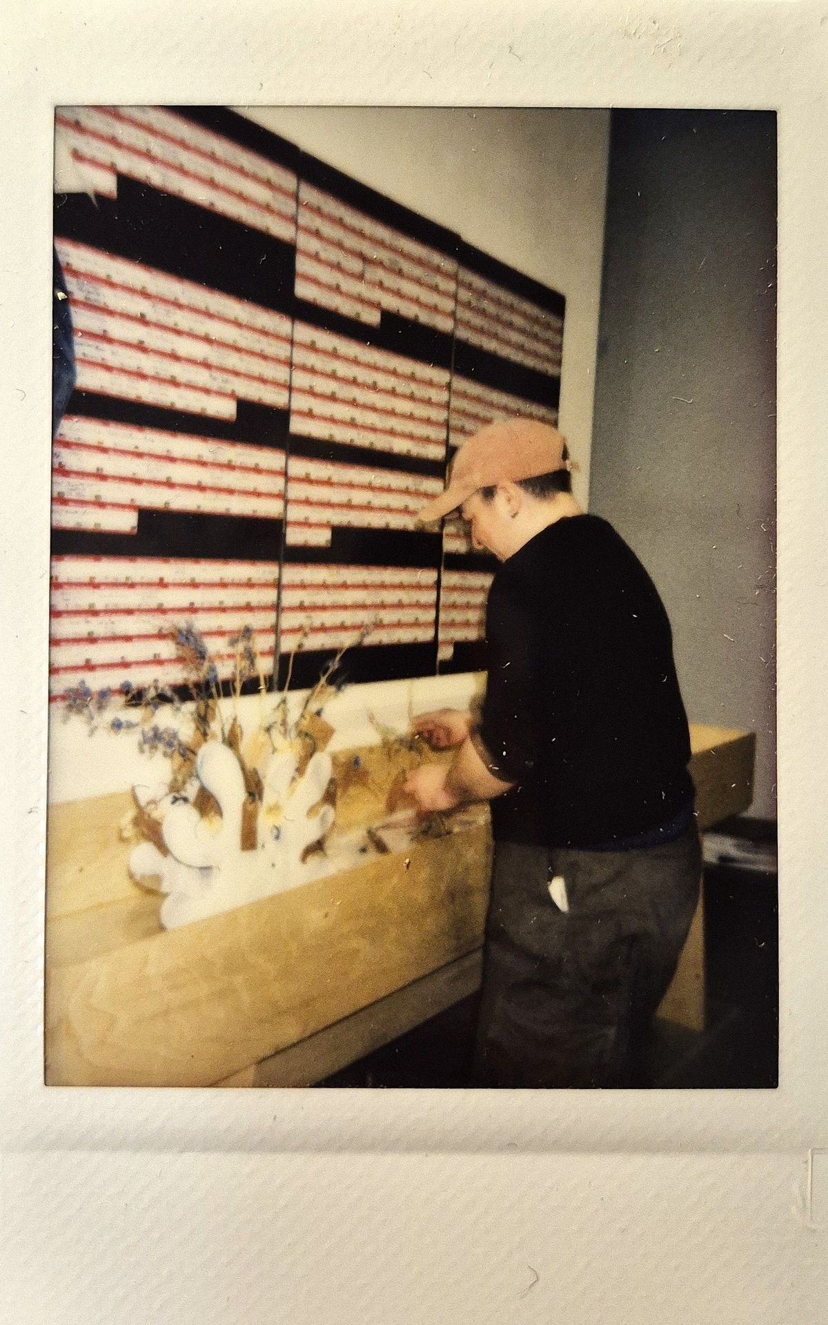 A person wearing a cap is arranging dried flowers on a wooden table in front of a patterned wall.