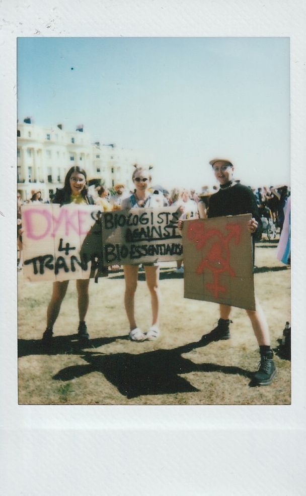 Three people stand with protest signs, gathered for a demonstration outside on a sunny day.