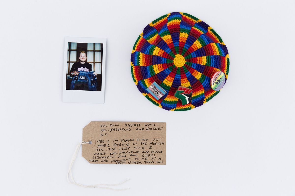 A rainbow coloured Kippah decorated with pro-palestine, pro-refugee, queer and trans pin badges. With a polaroid of the owner and a brown handwritten luggage tag.