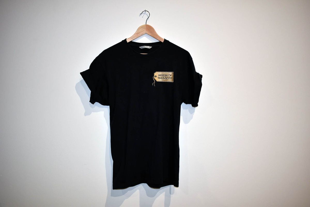 A black T-shirt with rolled sleeves which has the Museum of Transology logo on it hangs on a wooden hanger against a plain wall.