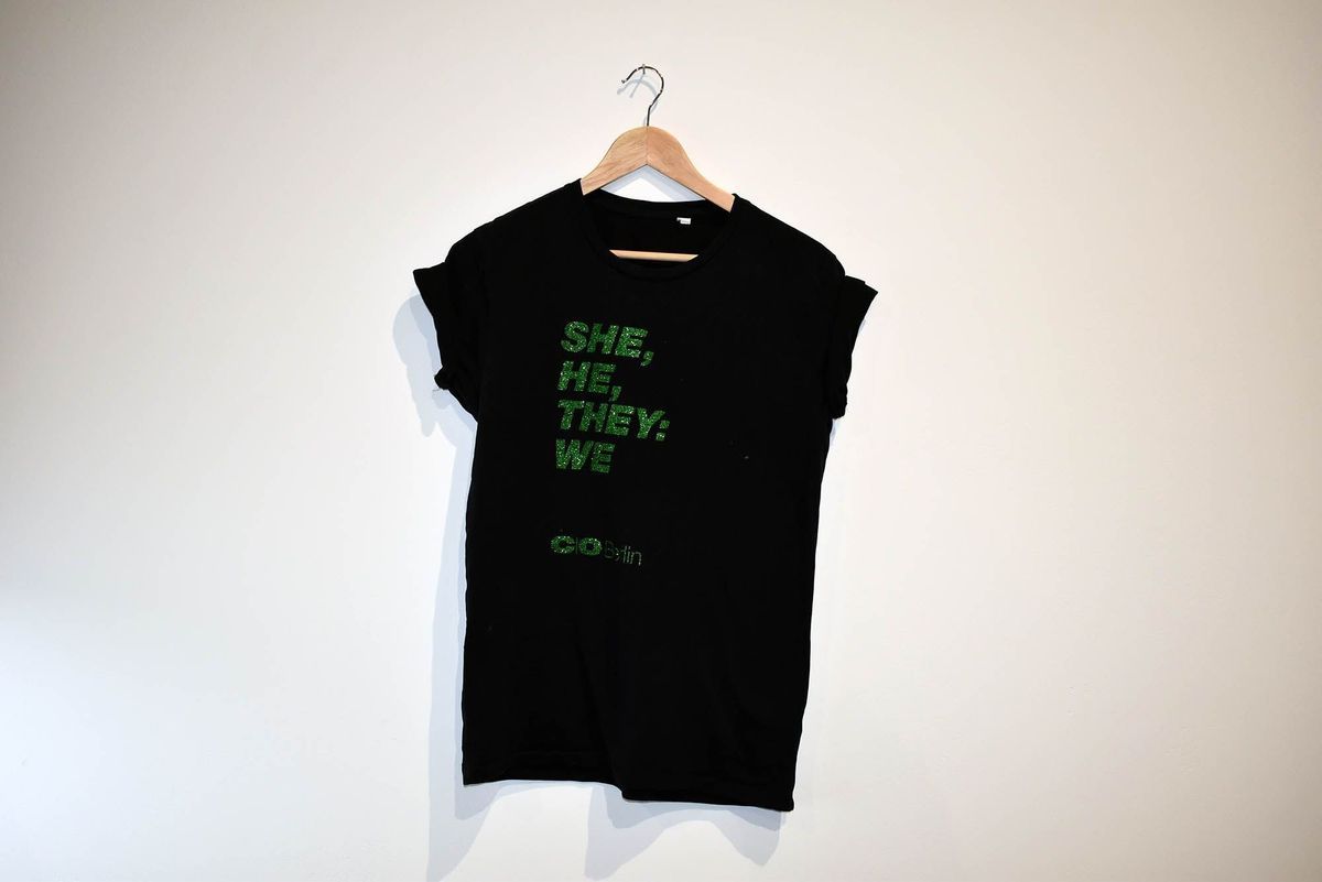 A black T-shirt on a hanger features green text which says 'She, He, They: We'.
