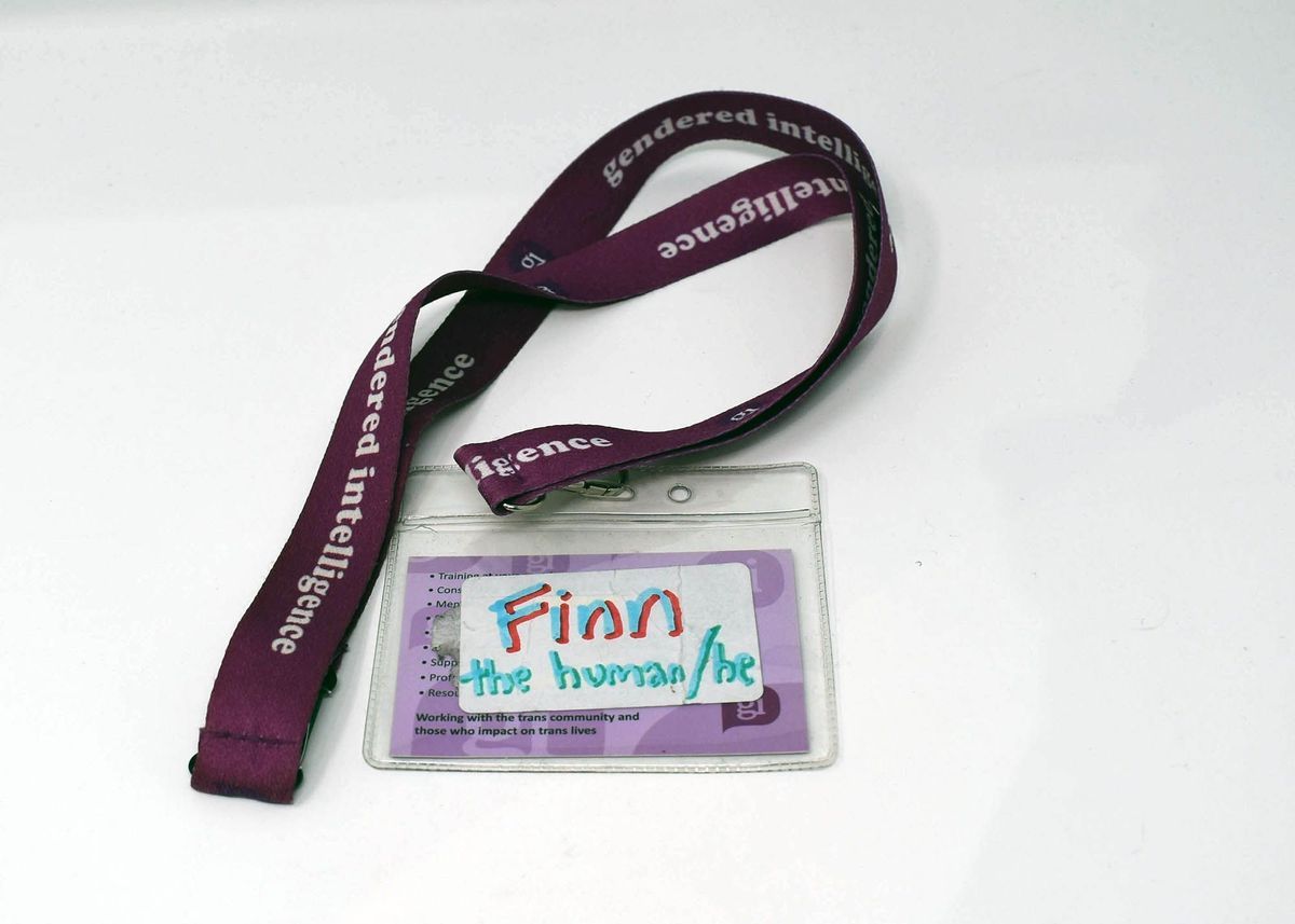 "Gendered intelligence" purple lanyard (470mm length) with plastic card holder, this contains a business card for "Finn Greig". The reverse has a sticker on it that says "Finn the human/he" in felt pen. Finn is lead youth worker at G.I.