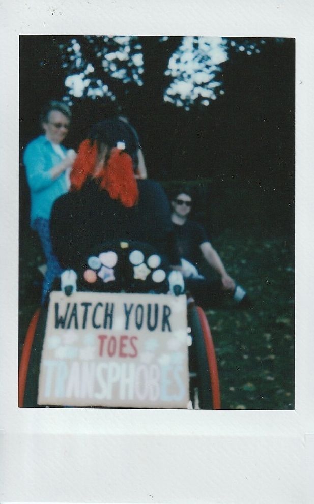 The back of someone sat in a wheelchair and there is a protest sign attached to the wheelchair which says 'Watch your toes transphobes'.