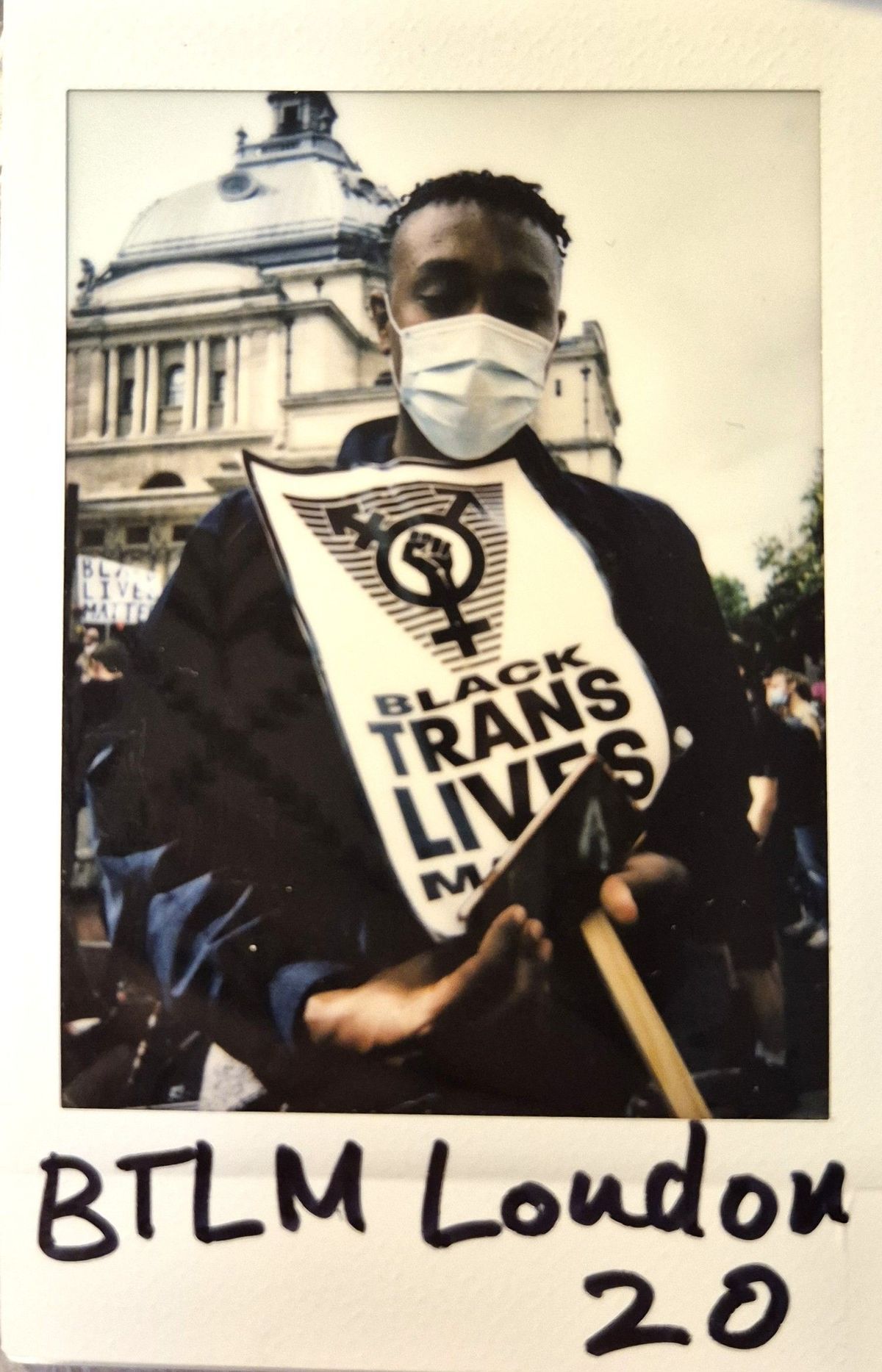 A person wearing a mask holds a "Black Trans Lives Matter" sign in front of a historic building