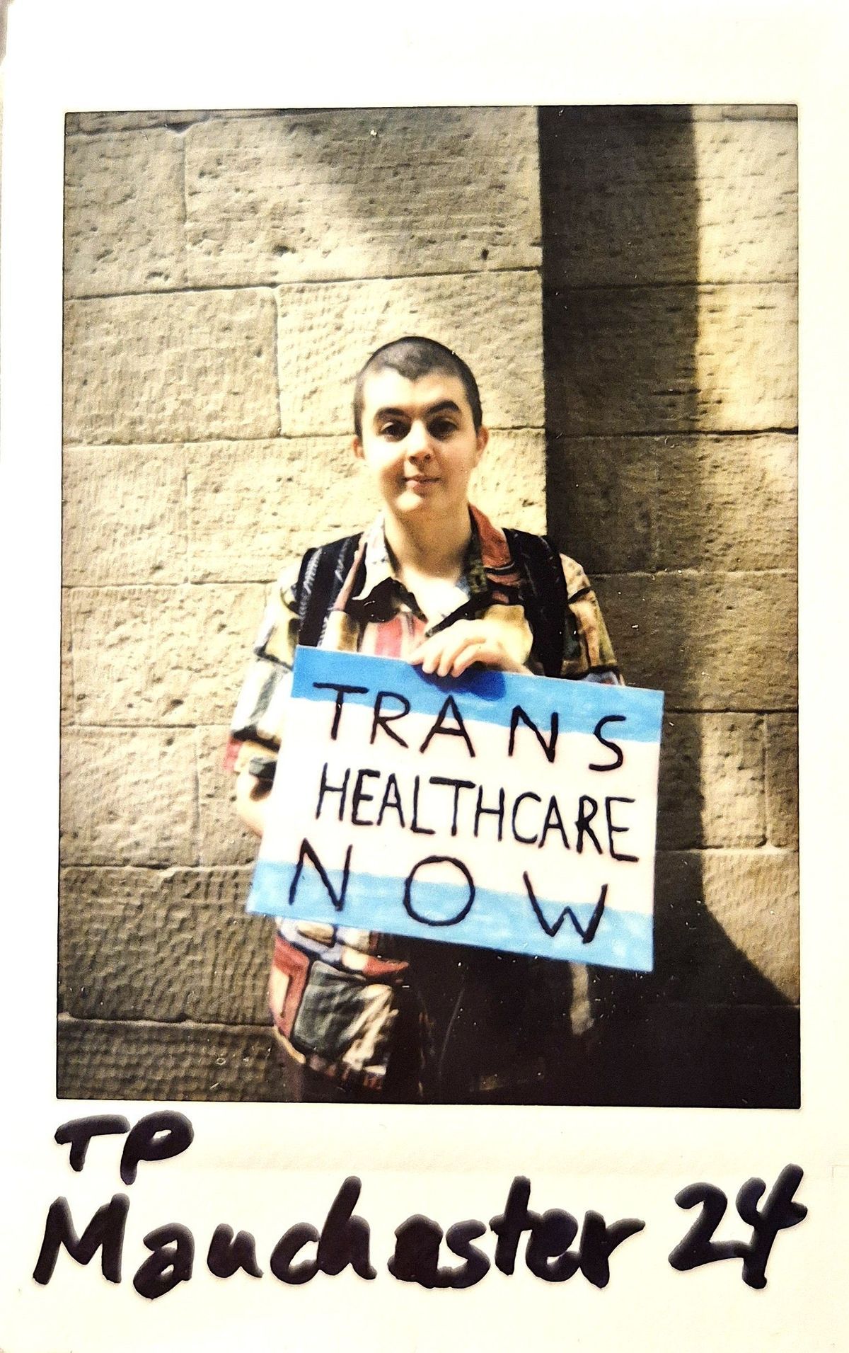 A polaroid of a person holding a sign reading "TRANS HEALTHCARE NOW" stands against a stone wall.