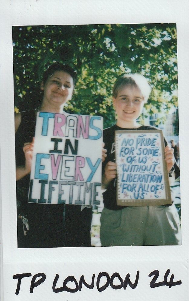 Two people hold signs promoting trans rights and pride, standing outdoors with trees in the background.