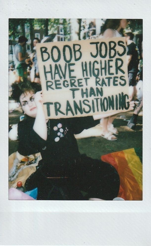 A person is sitting outdoors, holding a sign which says "BOOB JOBS HAVE HIGHER REGRET RATES THAN TRANSITIONING".