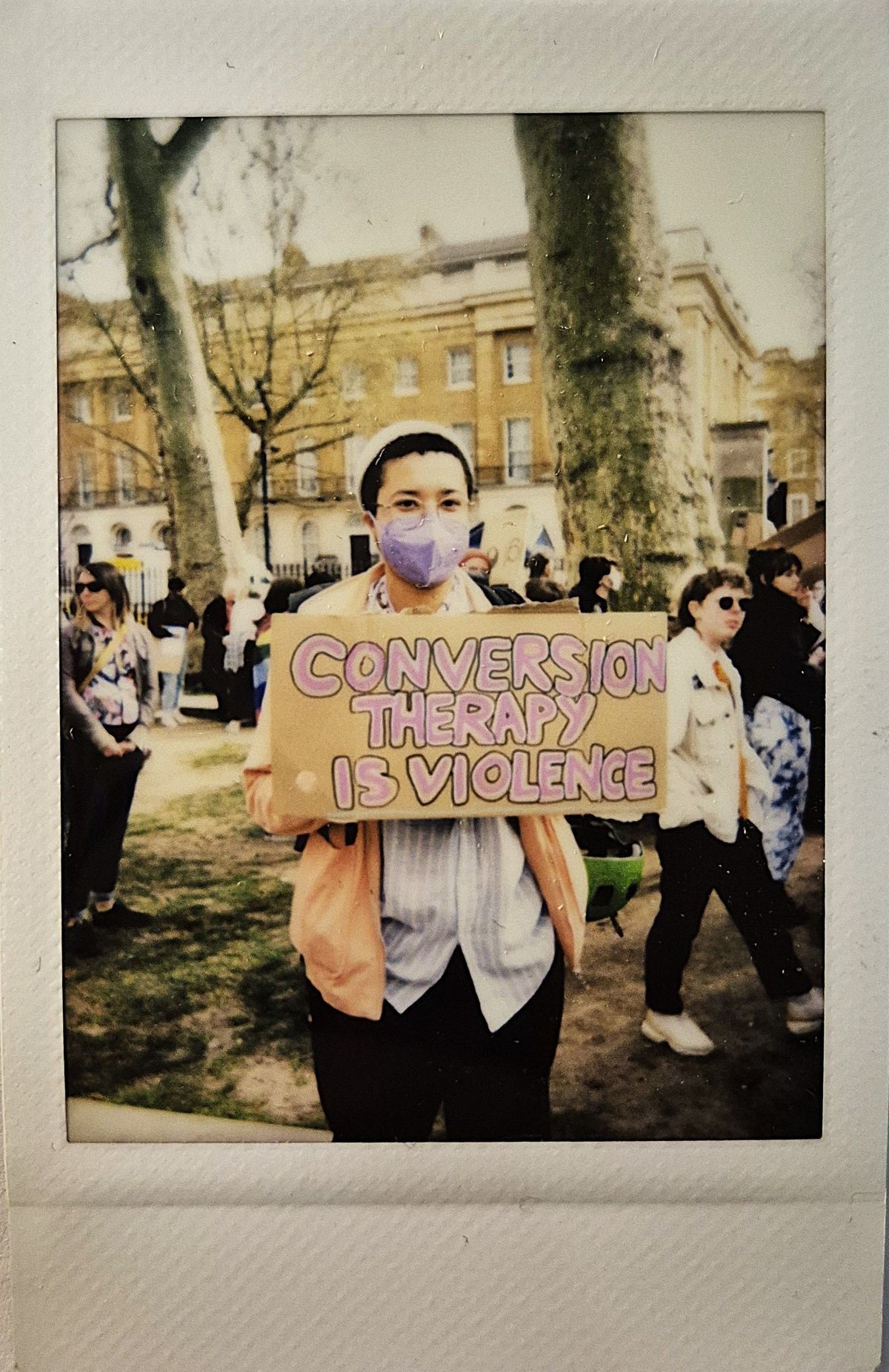 A person in a mask holds a sign saying "Conversion Therapy is Violence" during an outdoor protest.