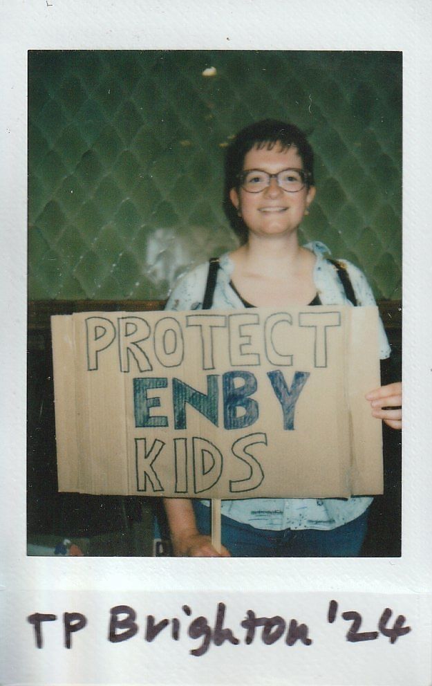 A person is holding a cardboard sign reading "PROTECT ENBY KIDS" with a smile.