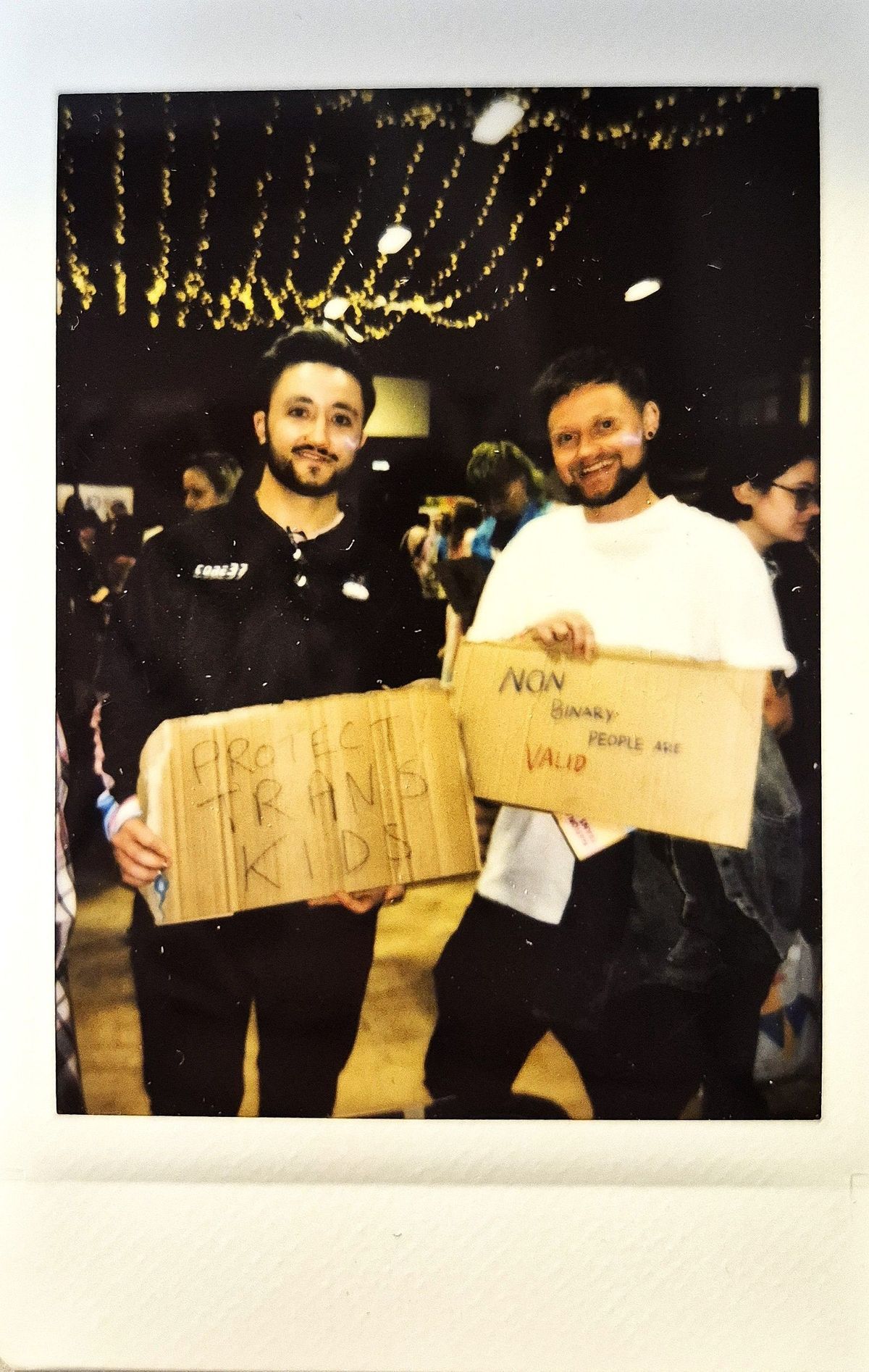 Two people hold signs advocating for trans children and non-binary individuals at an indoor event.