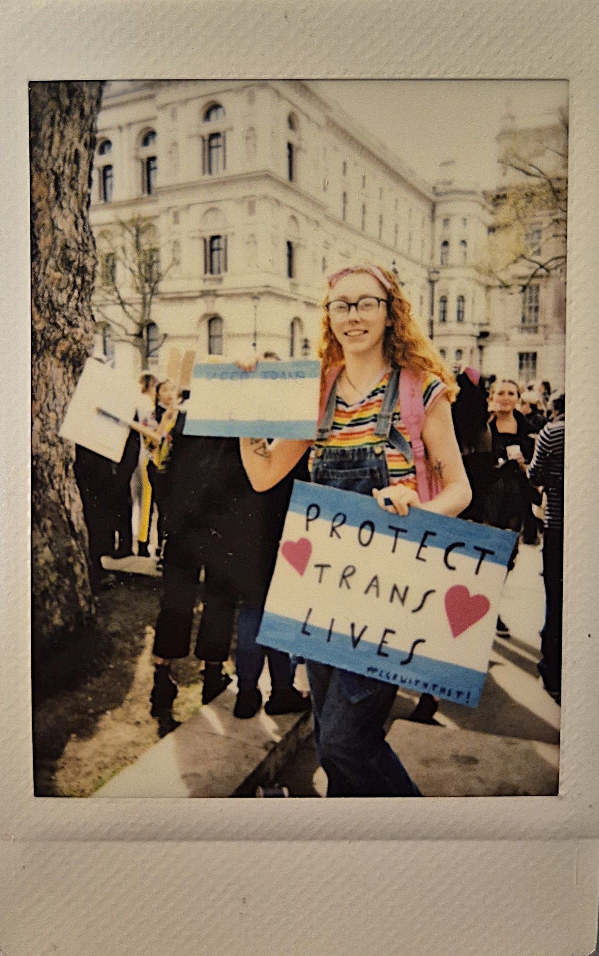 A person wearing colorful clothing holds a "Protect Trans Lives" sign at a rally.