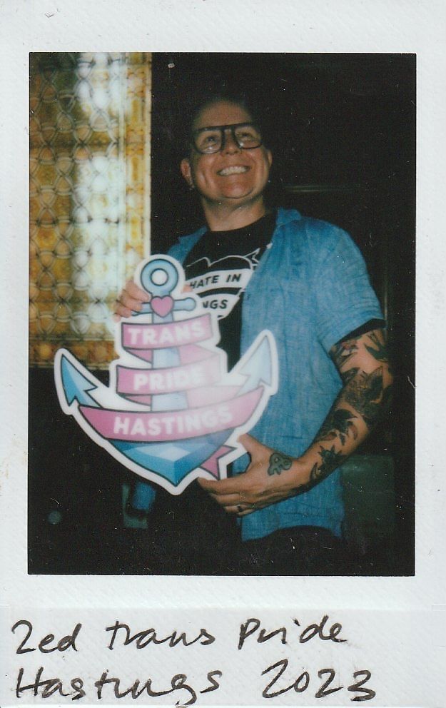 A person smiles while holding a sign that reads "Trans Pride Hastings,".