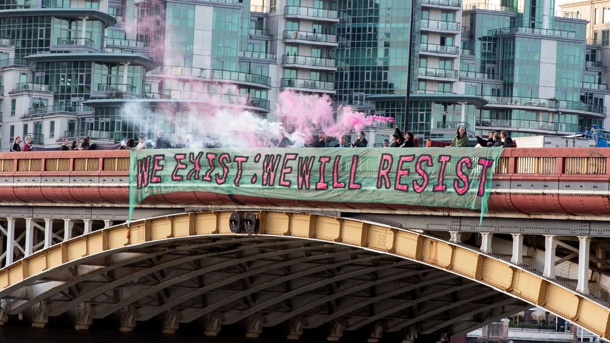 A banner which says 'We Exist: We Will Resist' and is a turquoise colour with the text in pink. It is being hung off a bridge in London with lots of people holding it with others letting off pink smoke. You can see London in the background built up with buildings.