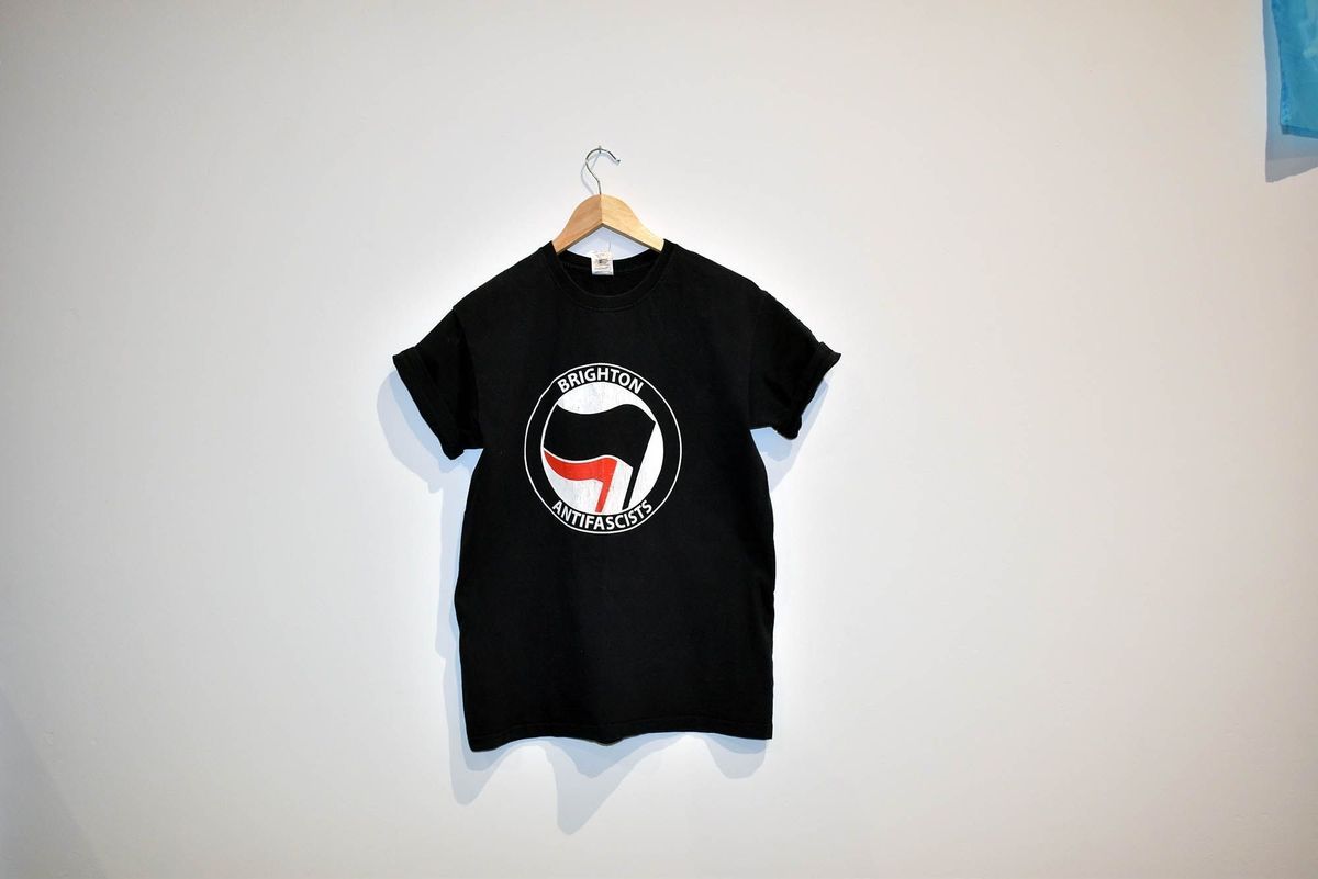 A black t-shirt with the "Brighton Antifascists" logo hangs on a wooden hanger.