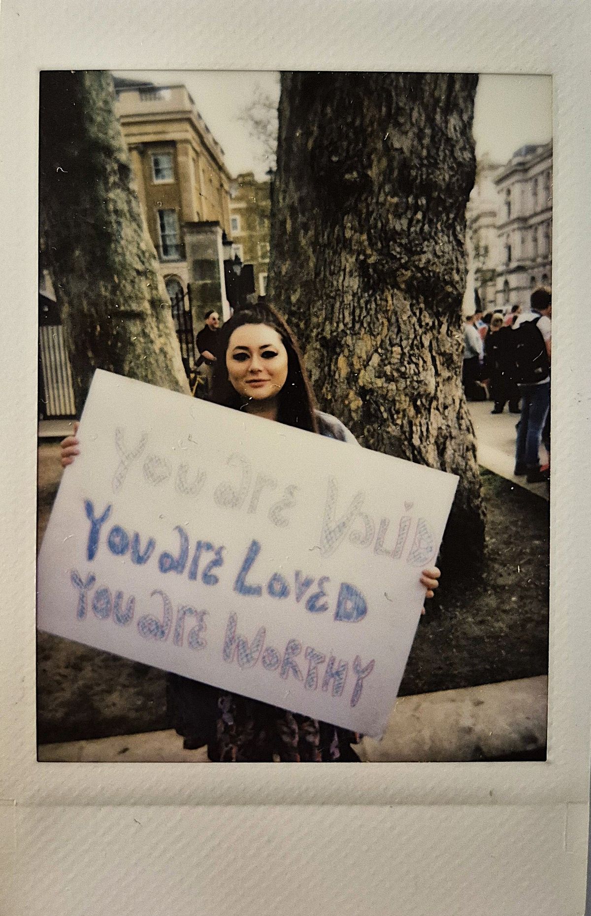 A woman stands outdoors holding a sign that reads, "You are valid, you are loved, you are worthy".