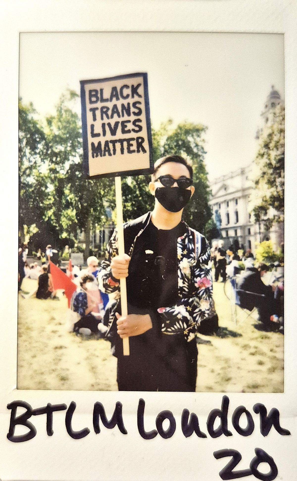 A person holding a "Black Trans Lives Matter" sign stands outdoors, wearing sunglasses and a mask.