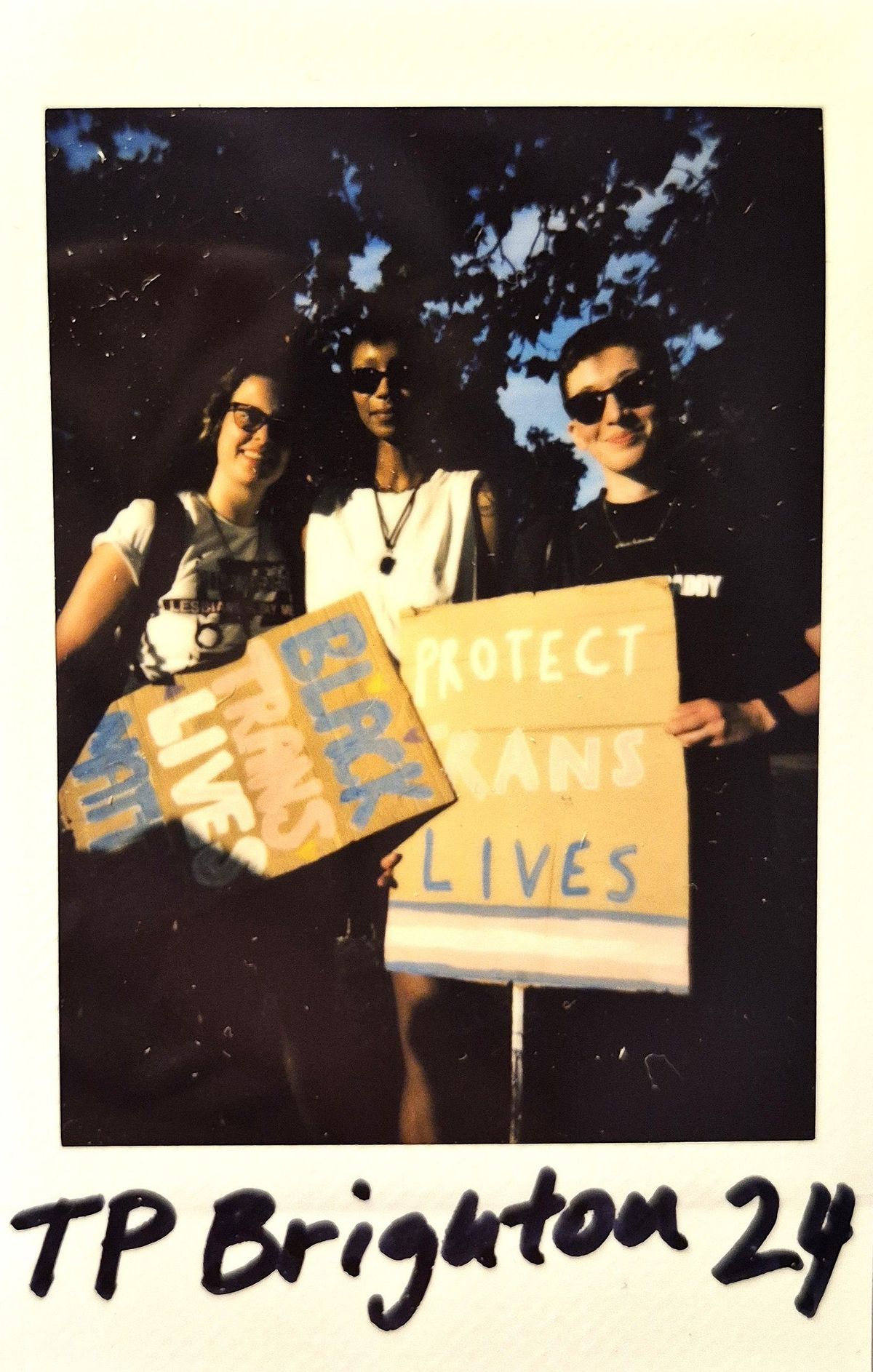 Three individuals are holding signs advocating for Black trans lives, standing outdoors under a tree.