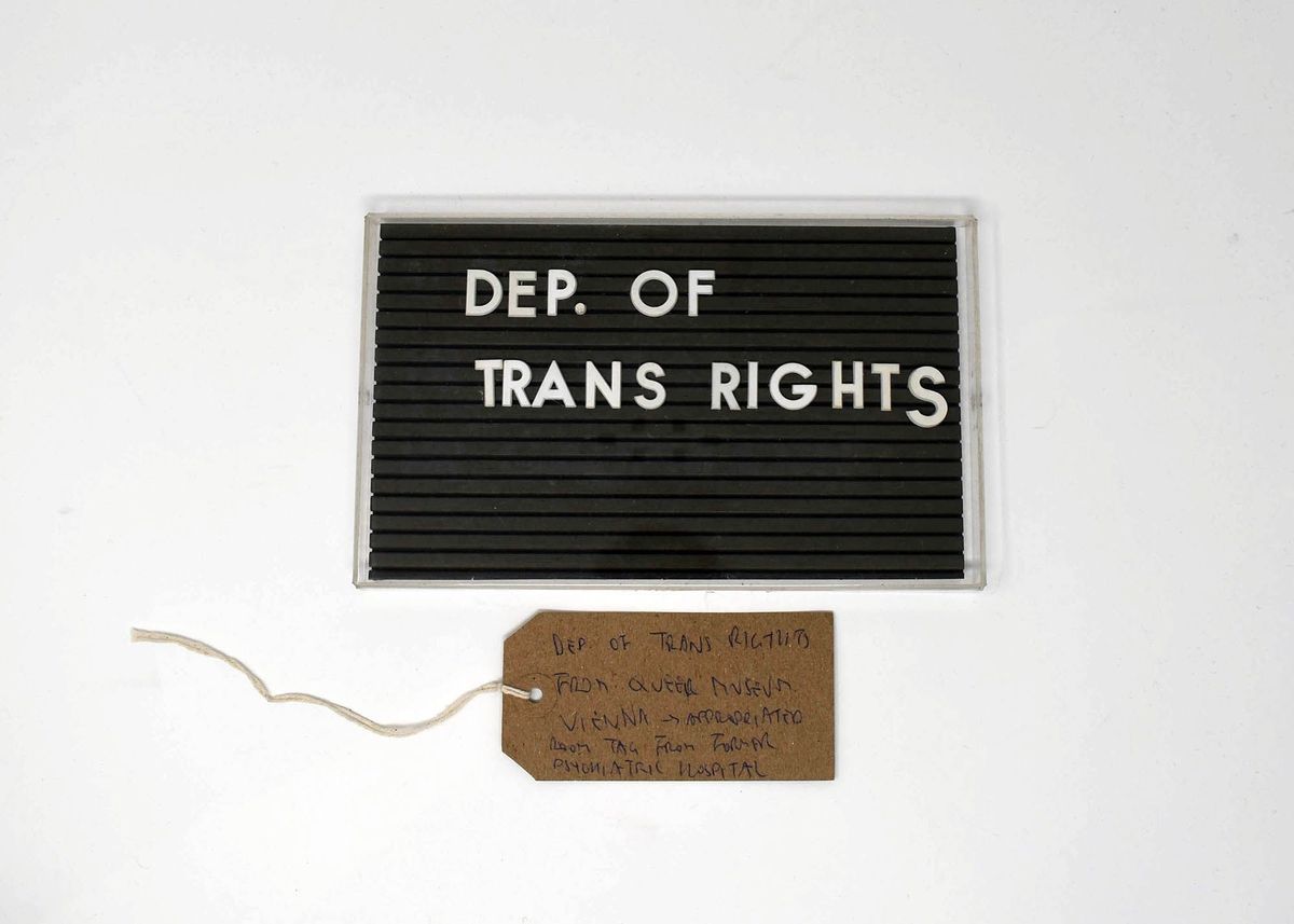 A black Marquee style sign board with the lettering "DEP. OF TRANS RIGHTS" in white letters. With a brown, handwritten luggage tag.
