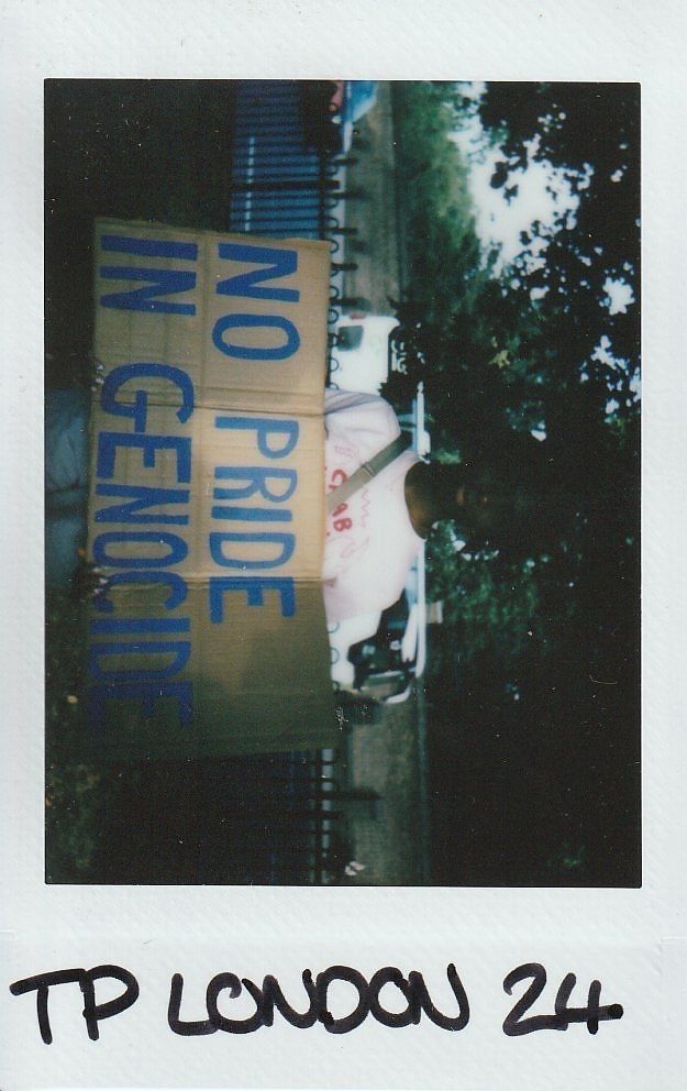A person holds a cardboard sign with "No Pride in Genocide" written in bold blue letters.