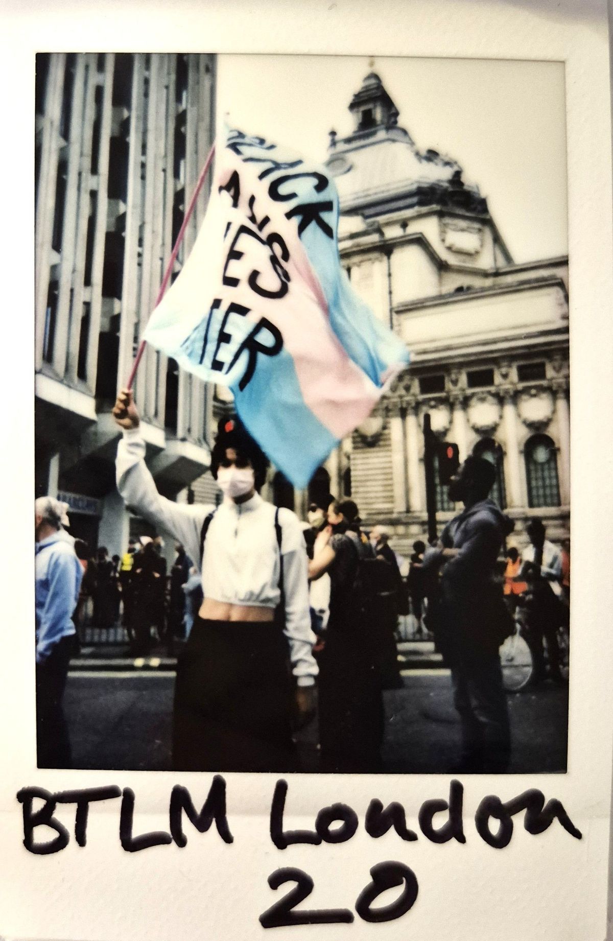 A person wearing a mask holds a trans flag during a protest in London, with buildings visible behind.
