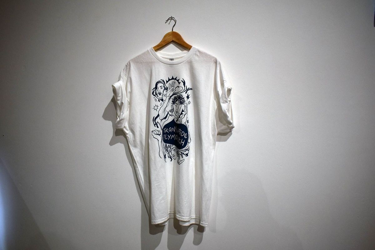 A white T-shirt with an artistic design hangs against a plain wall background.