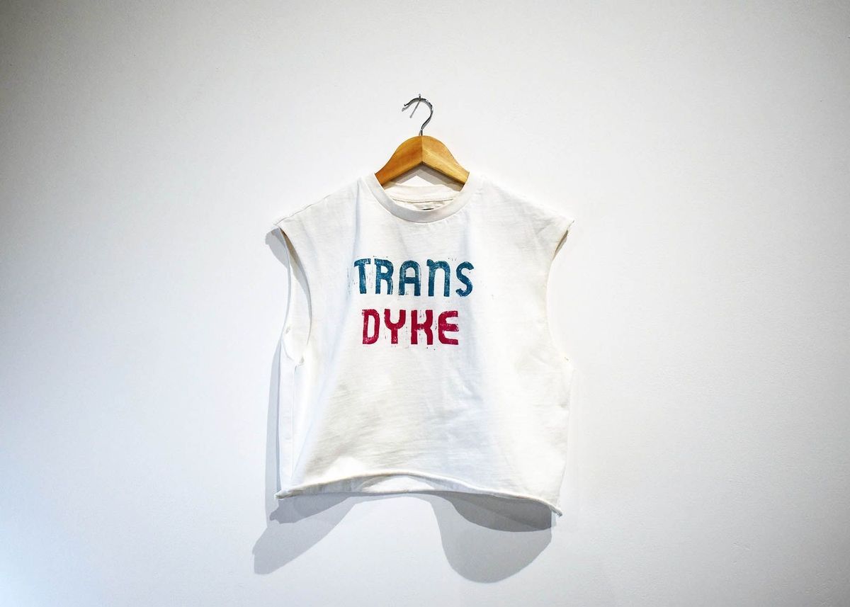 A white sleeveless shirt hangs on a wooden hanger, with Trans Dyke on it.