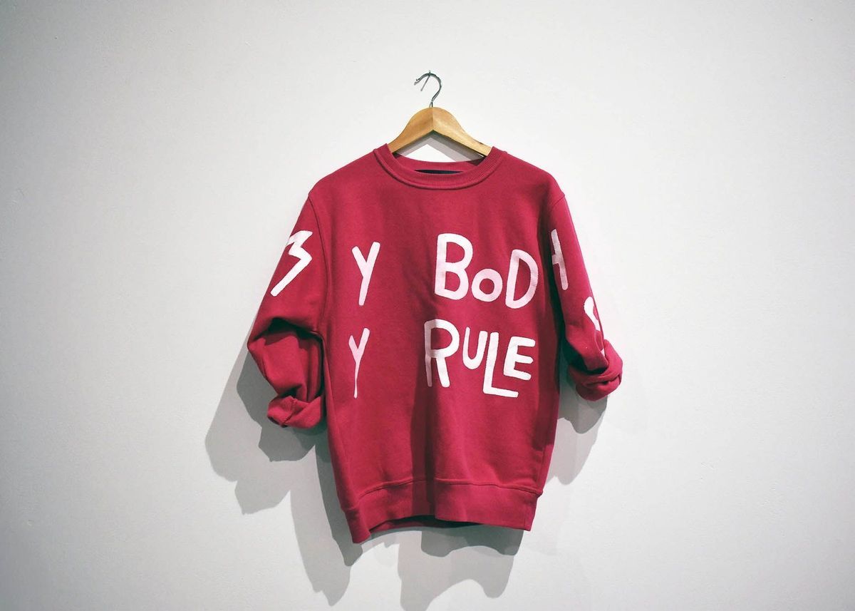A red sweatshirt with white text hangs on a wooden hanger against a plain wall.