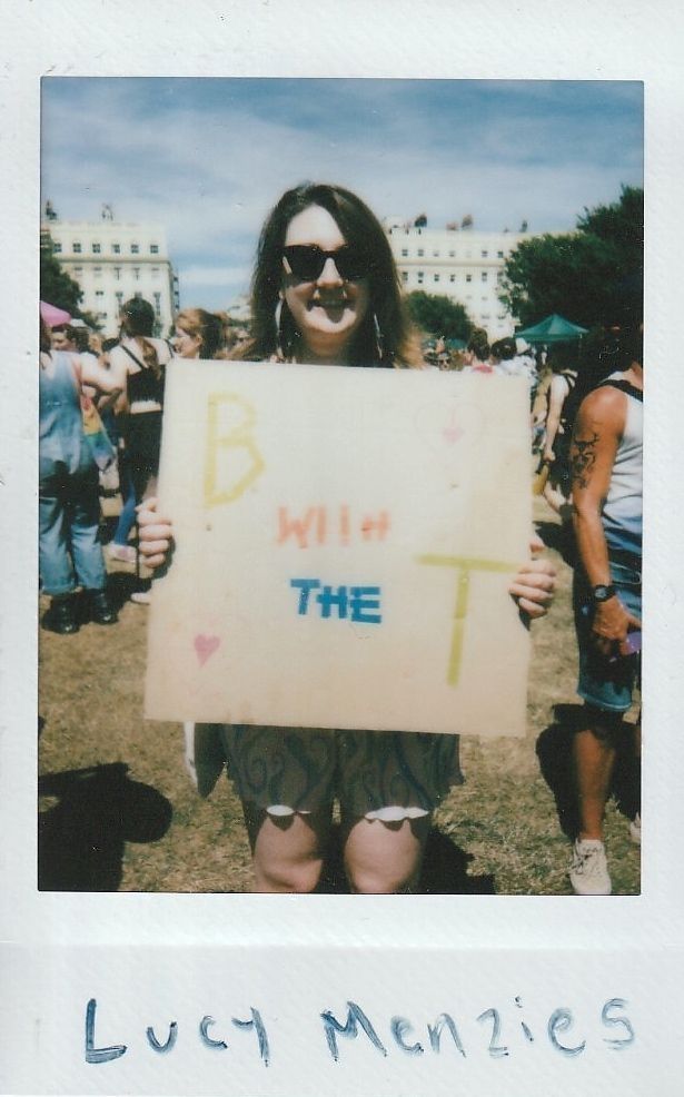 A person at an outdoor event holds a sign reading "B with the T" while smiling.