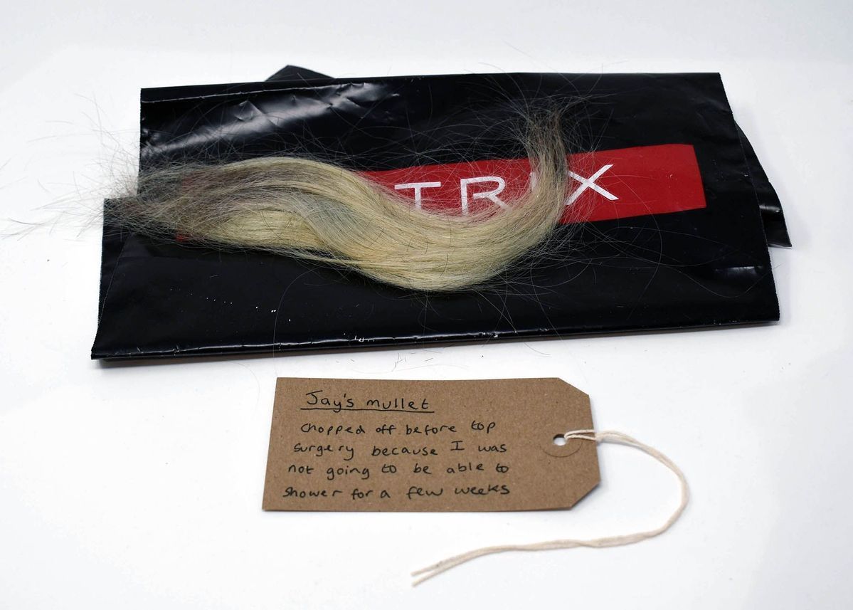 A section of blonde hair lays on a black plastic bag with the text "MATRIX" in white on a red background. With a brown, handwritten luggage tag.