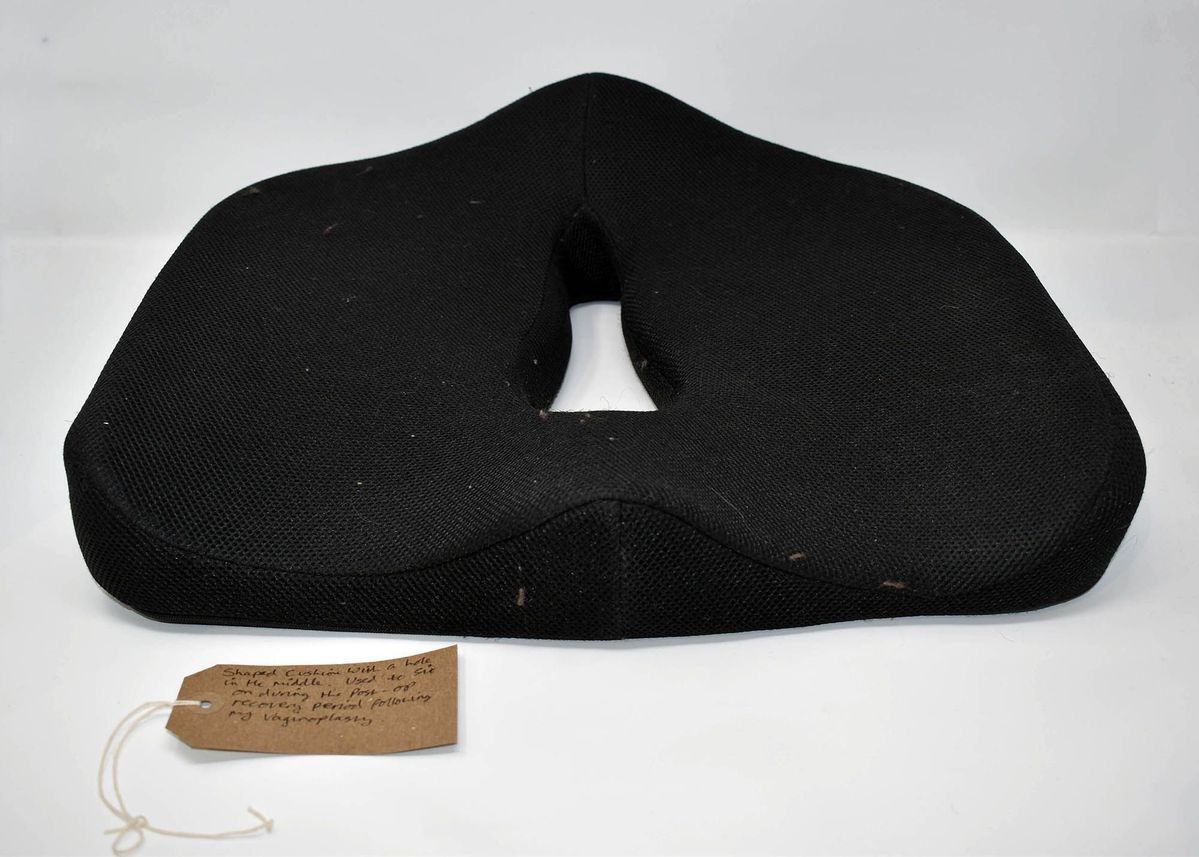 A black, shaped donut cushion for use after vaginoplasty surgery. With a brown, handwritten luggage tag.