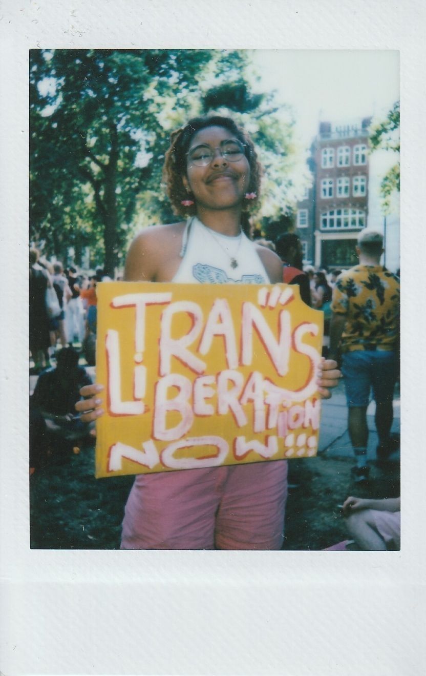 A person holds a vibrant sign reading "Trans Liberation Now" at an outdoor gathering under trees.