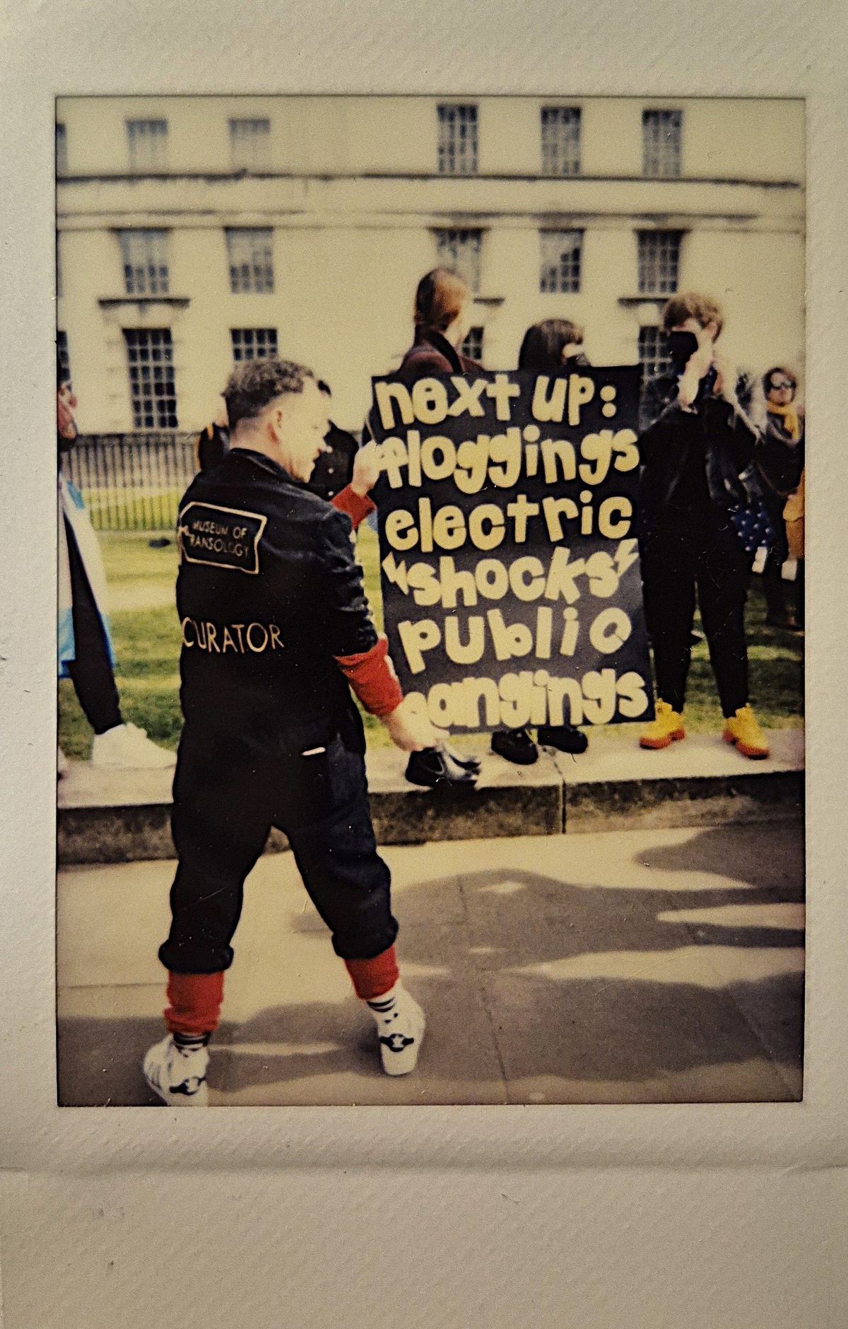 A person stands in front of individuals holding a handmade sign which says "NEXT UP: FLOGGINGS ELECTRIC SHOCKS PUBLIC HANGINGS".