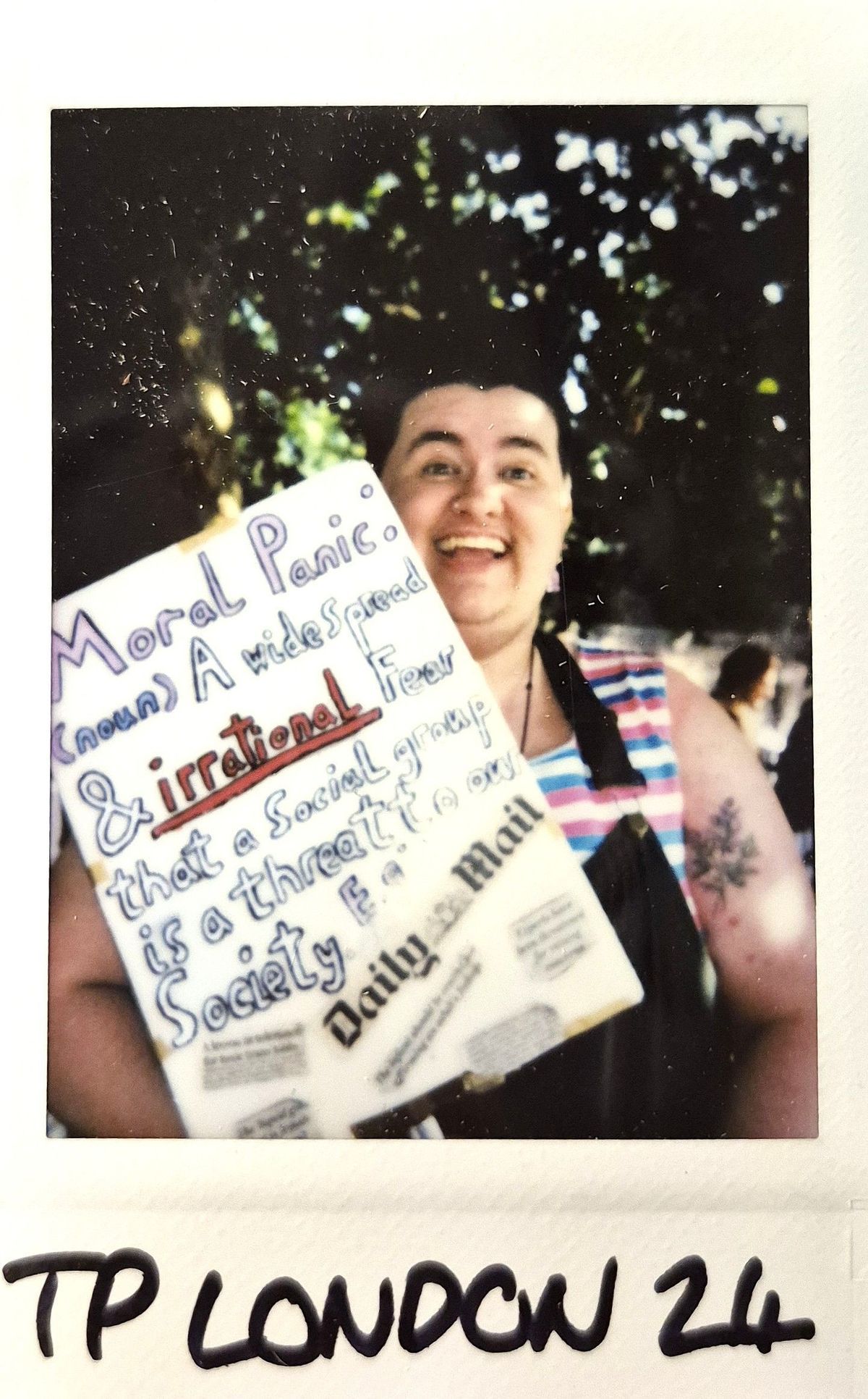 A person holds a colorful sign about moral panic, smiling in a sunny outdoor setting.