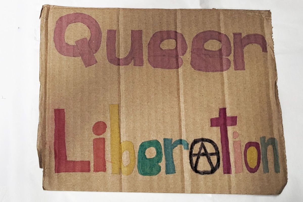 The image features a cardboard sign stating "Queer Liberation".