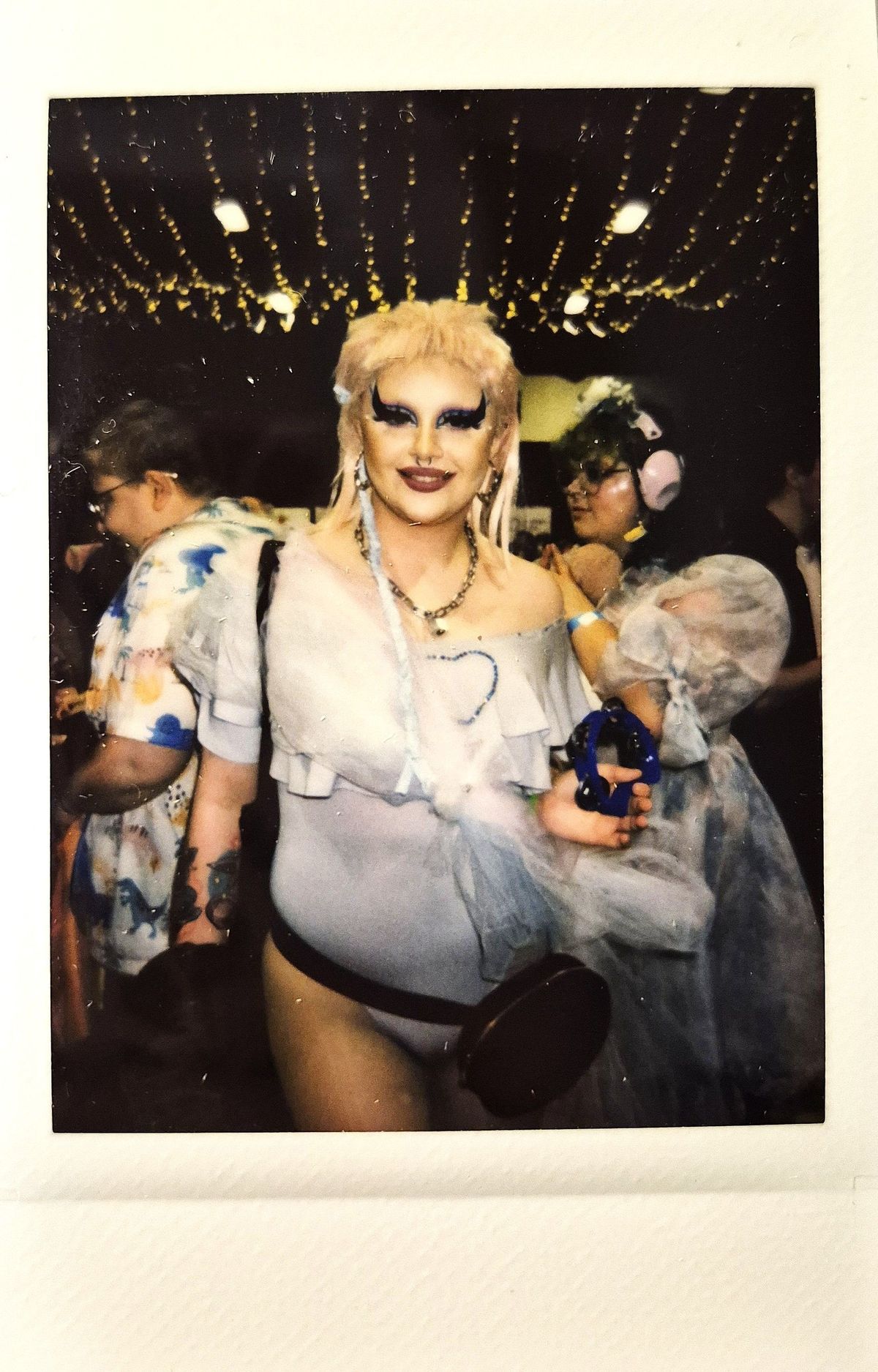 A person in elaborate makeup, wearing a stylish outfit, holds a tambourine in a festive space.