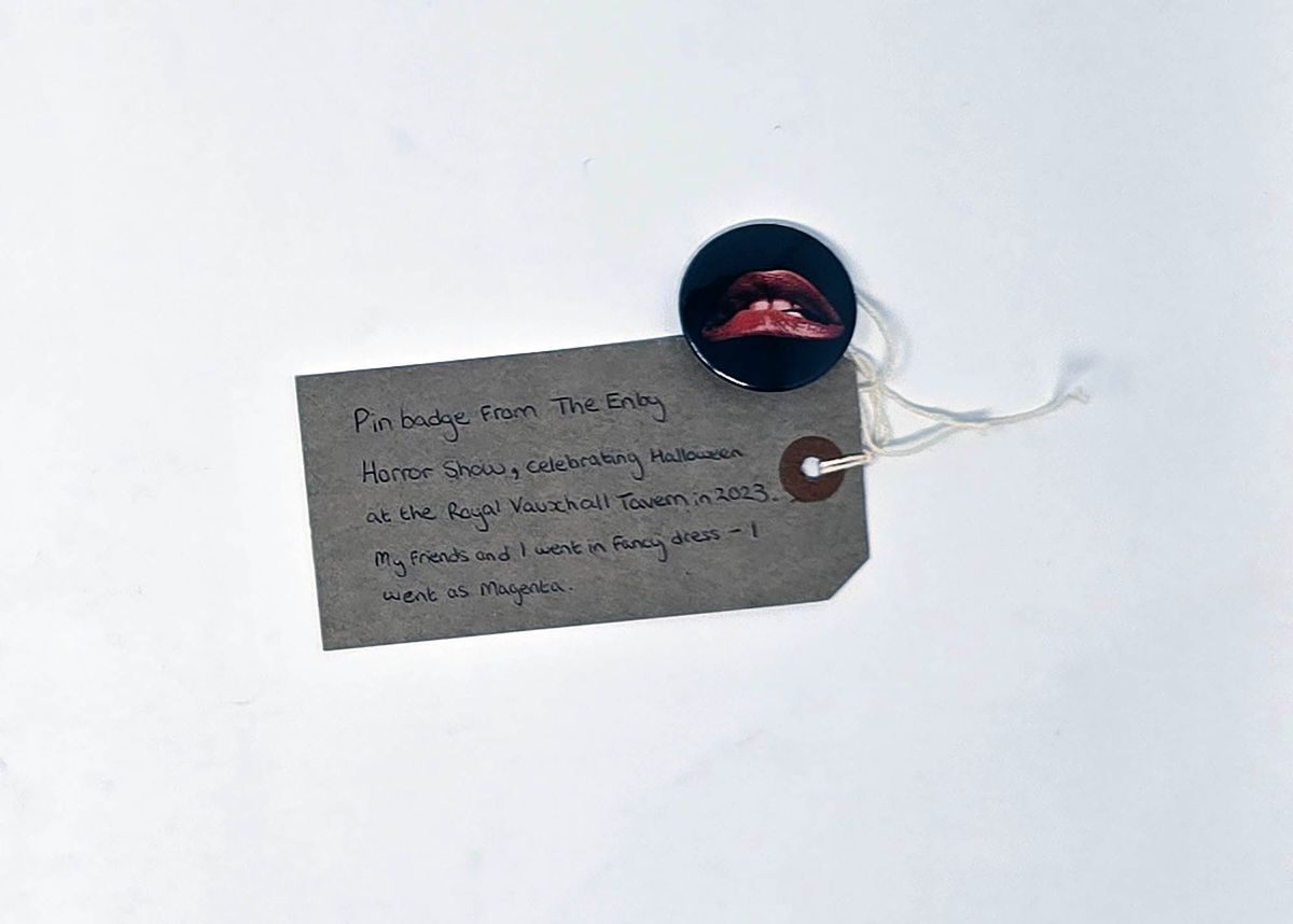 A small badge featuring red lips on a black background. The image is synonymous with the Rocky Horror Picture Show. With a brown, handwritten luggage tag.