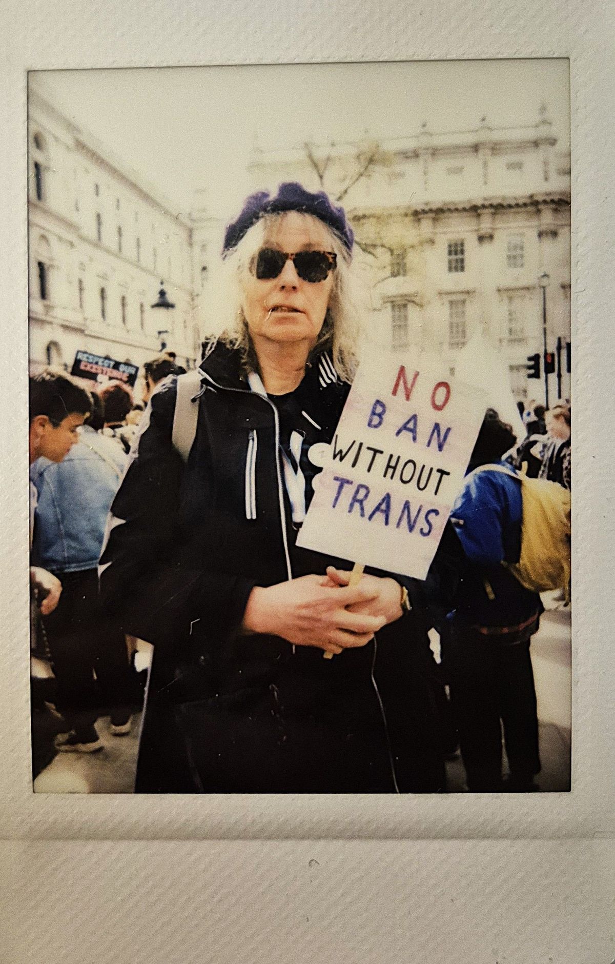 A person wearing sunglasses holds a sign reading, "NO BAN WITHOUT TRANS," at a protest.